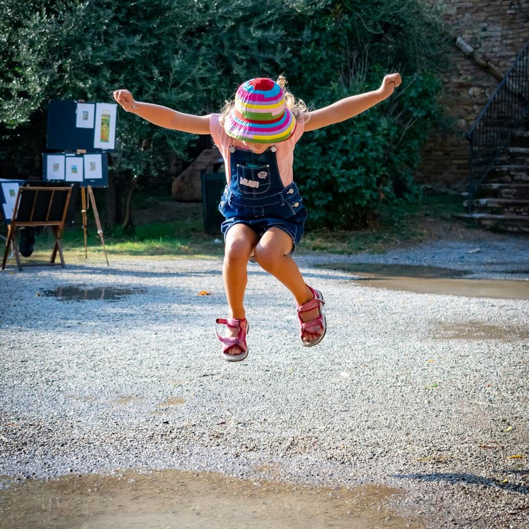 an image of a child with a brightly colored hat, jumper, and sandals, jumping with their arms spread wide.